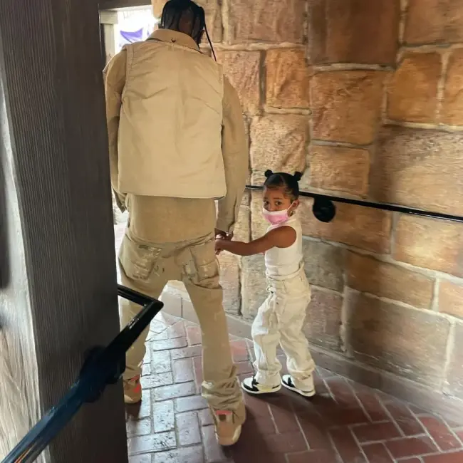 Travis Scott ve kızı Stormi'nin, aynı tarzda, açık renk kıyafetler ve spor ayakkabılar giyerek uyumlu bir şekilde yürüdükleri bir iç mekan çekimi.