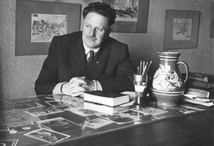 Nazım Hikmet, bir masanın başında ellerini kenetlemiş otururken; etrafında kitaplar, kalemlik ve sanatsal objeler yer alıyor.