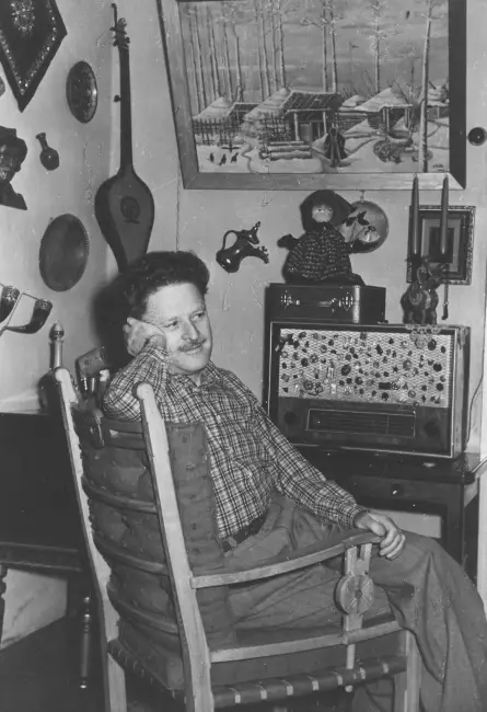 Nazım Hikmet, oturma odasında radyo başında düşünceli bir ifadeyle poz veriyor; duvarda geleneksel objeler asılı.