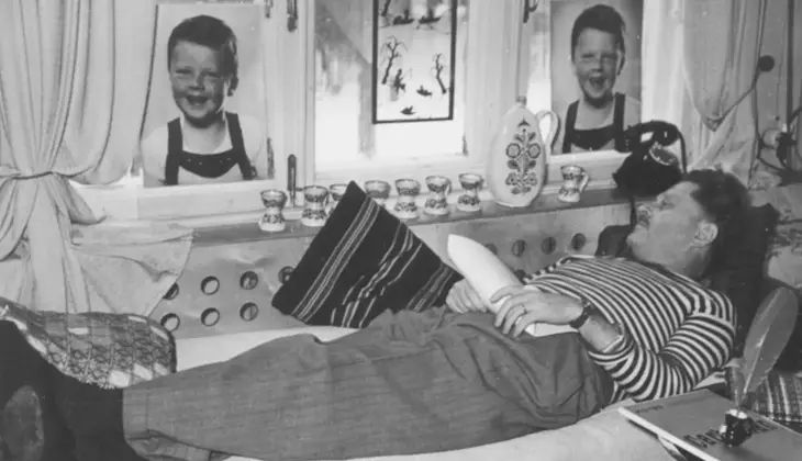 Nazım Hikmet, ev ortamında pencerede çocuk fotoğrafları önünde dinlenirken, elinde kâğıtla uzanıyor.