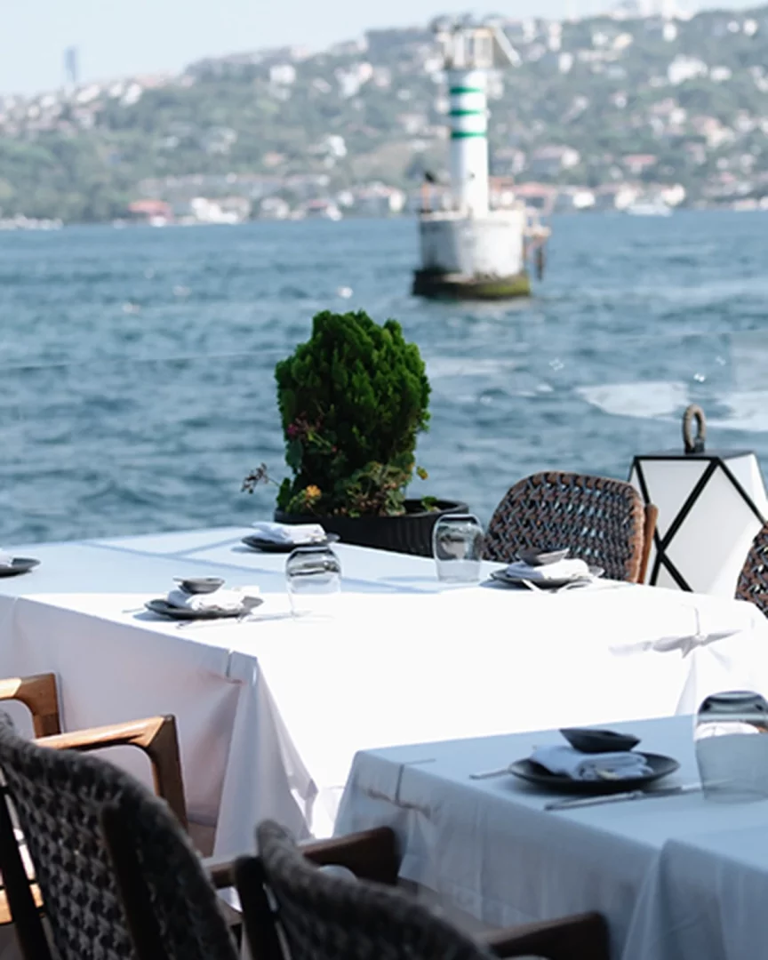 İstanbul Boğazı kıyısında, zarif beyaz masa örtüleriyle donatılmış yemek masaları ve hasır sandalyelerin ön planda olduğu şık bir restoranda, arka planda yeşil şeritli bir deniz feneri ve şehrin büyüleyici silüeti eşliğinde sunulan eşsiz bir Boğaz manzarası.