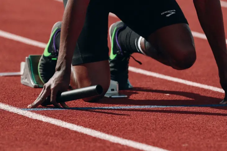 Kırmızı atletizm pistinde, beyaz başlangıç çizgisinin hemen gerisinde, siyah şortlu ve yeşil-mor detaylı spor ayakkabılı bir atletin alçak başlangıç pozisyonunda sağ eliyle bir bayrak çubuğunu tutarak beklediği yakın plan bir görsel, tüm zamanların efsaneleşmiş Olimpiyat sporcularının başlangıç anındaki kararlılığını ve rekabet ruhunu vurguluyor.