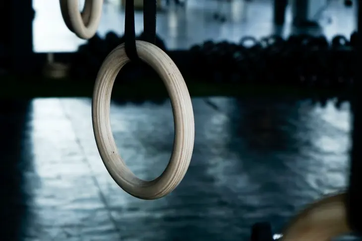 Koyu ve bulanık bir arka plan önünde asılı duran, pürüzsüz ahşap dokulu jimnastik halkaları, artistik jimnastik branşındaki efsaneleşmiş Olimpiyat sporcularının başarılarına ve zorlu mücadelesine işaret ediyor.