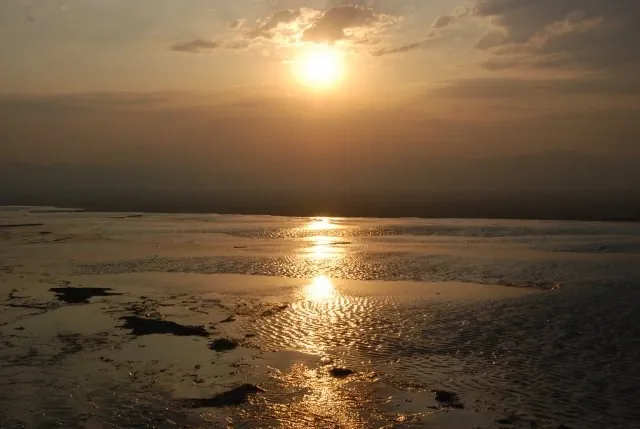 Güneşin batışıyla Pamukkale travertenlerinin sığ, teraslı havuzlarında altın rengi ışıkların suya yansıyarak parıldadığı, ufukta dağ silüetlerinin belirdiği dingin ve büyüleyici bir manzara.