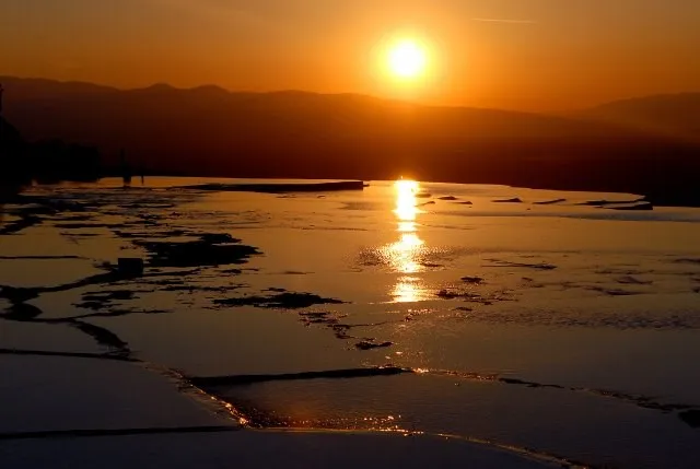 Pamukkale Travertenleri üzerinde gün batımının altın rengi ışıklarıyla yıkanan bembeyaz teraslar, su birikintilerinde parlayan güneşin ve uzak dağların silüetleriyle adeta cennetten bir köşe sunarak büyüleyici bir doğa harikası manzarası oluşturuyor.