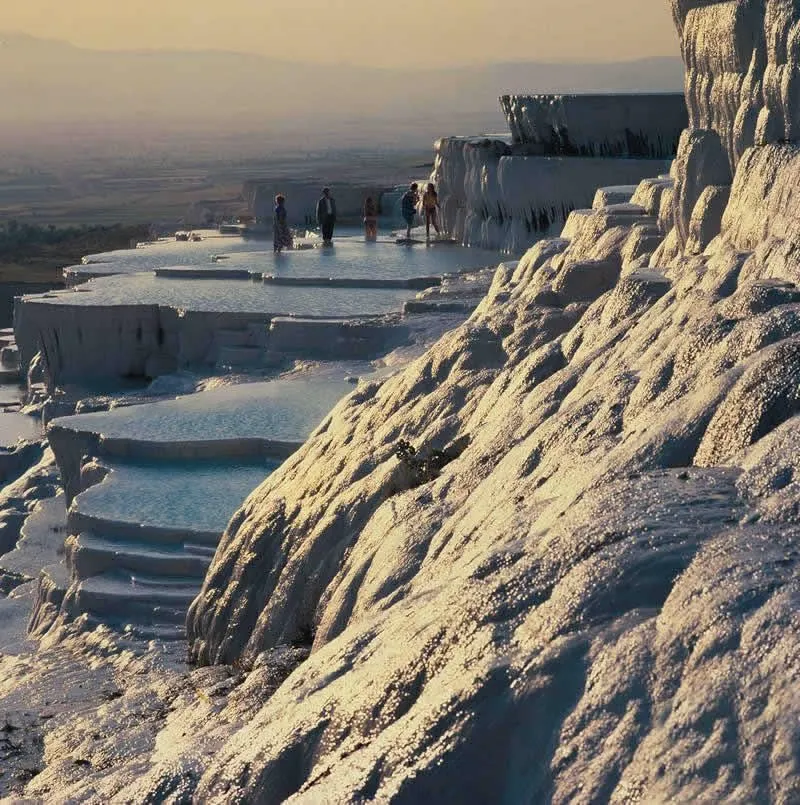 Pamukkale Travertenleri, gün batımının sıcak altın rengi ışıklarıyla aydınlanmış, kıvrımlı bembeyaz kireçtaşı yamaçlar ve berrak mavi suyla dolu katmanlı havuzlarda yalınayak yürüyen birkaç ziyaretçi ile doğanın büyüleyici bir anını yakalıyor.