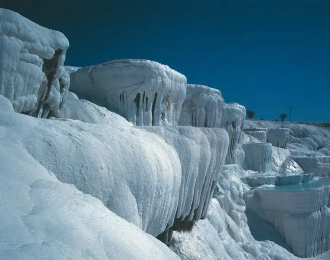Pamukkale travertenlerinin karakteristik bembeyaz, kademeli kireçtaşı terasları, sıcak termal suların oluşturduğu doğal ve pürüzlü yüzeyleriyle kadrajı doldururken, üst kısımdaki derin mavi gökyüzü bu etkileyici jeolojik yapıyı çarpıcı bir kontrastla vurguluyor ve bölgenin ikonik görünümünü özetliyor.