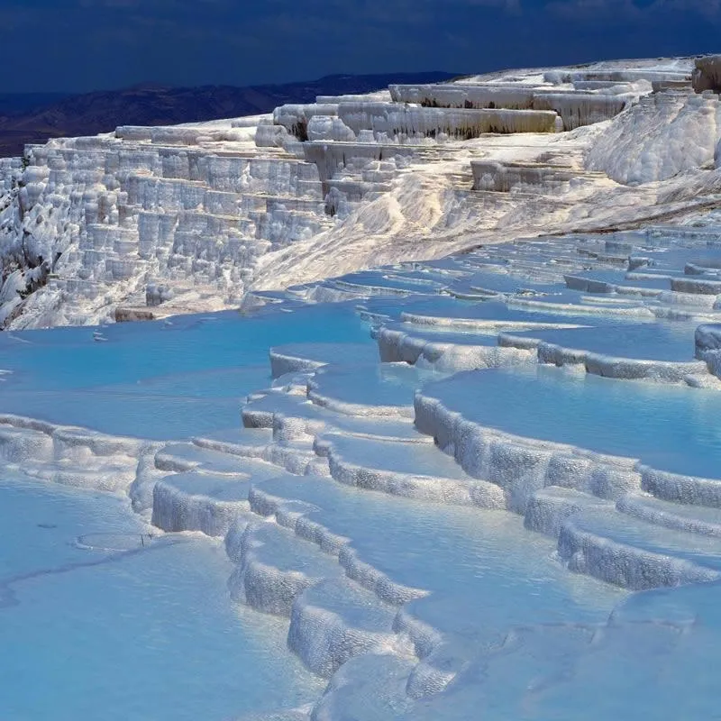 Pamukkale Travertenleri'nin, kireçtaşıyla bembeyaz pamuk tarlalarını andıran katmanlı terasları ve içlerini dolduran berrak turkuaz termal suları, uzakta yükselen koyu tepeler ve bulutlu gökyüzü altında doğanın benzersiz bir sanat eserini sergiliyor.