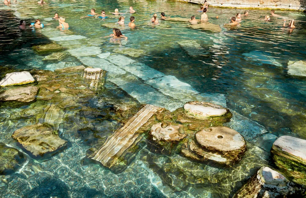 Pamukkale'nin antik termal havuzunda, berrak turkuaz sular içinde yüzen ziyaretçiler arasında, suyun derinliklerinde uzanan Hierapolis'ten kalma antik sütun ve taş kalıntıları açıkça seçiliyor.