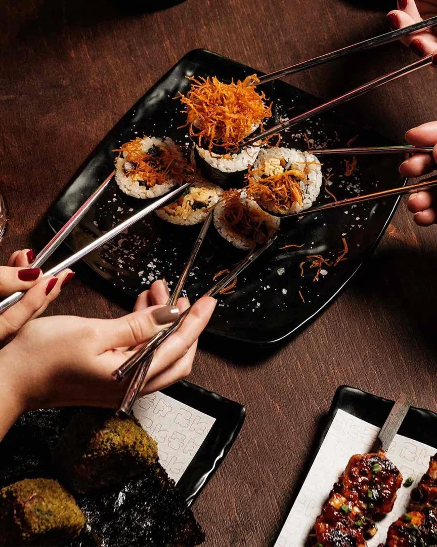 Çeşitli ellerin, parlak metal çubuklarla siyah dikdörtgen bir tabaktan üzerleri çıtır kızarmış tellerle zenginleştirilmiş sushi roll'ları alırken gösterildiği bu kare, ahşap bir masada paylaşımlı bir Japon yemeği deneyimini canlı bir şekilde yansıtmaktadır.