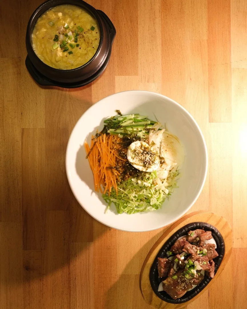 Seorabeol Kore Restoranı'nda ahşap bir masada servis edilen, havuç, lahana, salatalık, deniz yosunu ve susamla zenginleştirilmiş üzeri yumurtalı doyurucu bir bibimbap kasesinin yanında, taze soğanlı kremamsı bir yumurta yemeği ve bol soslu ızgara et tabağı görülüyor. İstanbul Uzak Doğu restoranları arasında yer alan Seorabeol Korean Restaurant mekânından bir görünüm.