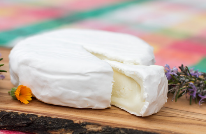 Edremit Körfezi'nin gastronomik zenginliklerini vurgulayan, İdamera Çiftliği'nin beyaz küflü, kesilmiş ve içi kremsi görünen koyun sütü Tamambert peyniri, ahşap bir sunum tahtasında mor ve sarı çiçeklerle süslenmiş.