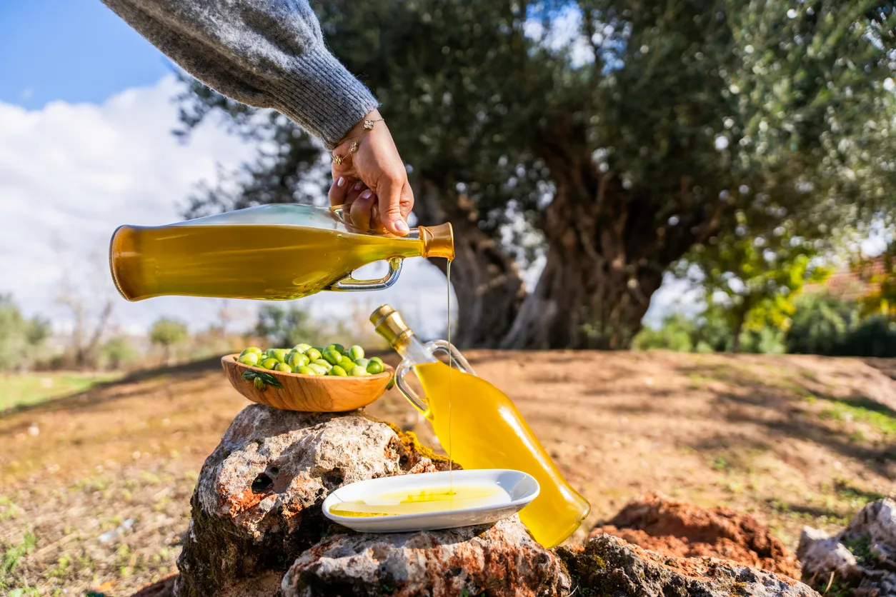 Bir el, açık havada, zeytin ağaçlarının yeşillikler içindeki doğal ortamında, bir şişeden altın sarısı zeytinyağını başka bir şişeye ve küçük bir tabağa akıtırken, yanında taze yeşil zeytinlerle dolu ahşap bir kase ve kayalar görülüyor; bu görsel yaz besinleri arasında zeytinyağının önemini ve doğallığını vurguluyor.