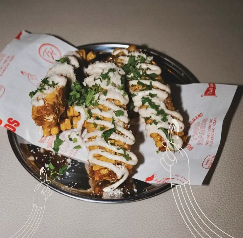 İzmir'in en iyi restoranlarından biri olan Two Hands Burger & Toastery'de, beyaz krema sosu, taze yeşillikler ve rendelenmiş parmesan peyniriyle zenginleştirilmiş, karamelize edilmiş ve hafifçe kızarmış lezzetli trüflü parmesanlı mısır kaburgaları parlak metal bir tabakta iştah açıcı bir şekilde sunuluyor.