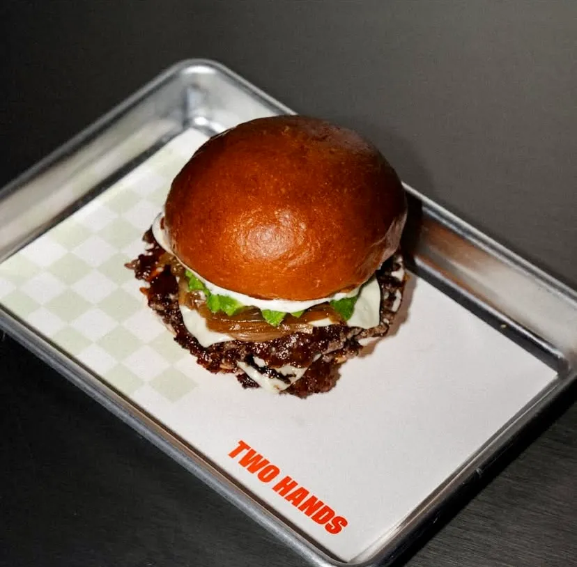 İzmir'in En İyi Restoranları başlığı altındaki makalede öne çıkan, Two Hands Burger & Toastery'ye ait, metal bir servis tepsisinde sunulan, parlak brioche ekmeği, bol et köftesi, erimiş peynir, karamelize soğan ve taze yeşilliklerle dolu, ağız sulandıran bir hamburger.
