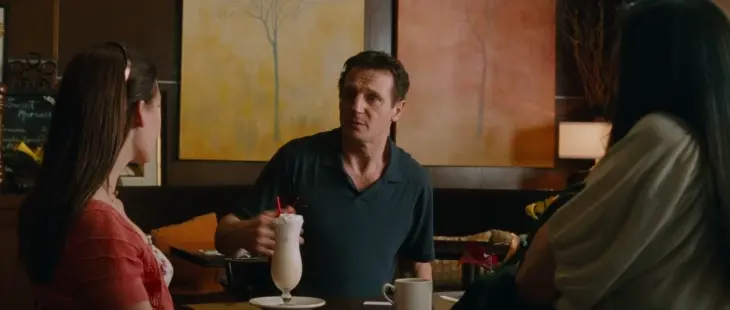 Taken filminden Bryan Mills (Liam Neeson), bir kafede kızına bakarken, endişeli ve kararlı bir ifadeyle.
