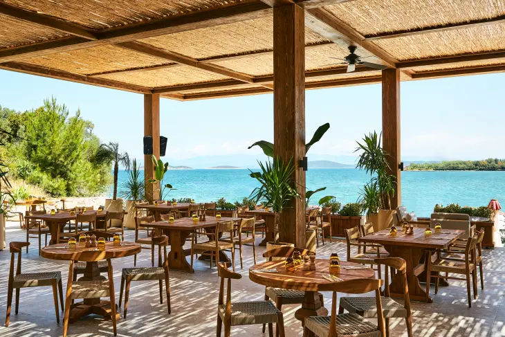 Bu fotoğraf, göz alıcı turkuaz denize bakan, hasır tavanlı ve ahşap direkli modern bir açık hava restoranın şık masalarını ve sandalyelerini gösteriyor; Bodrum'un en iyi otellerinden birinde unutulmaz bir yeme içme deneyimi vadeden doğal ve lüks atmosferi vurguluyor.