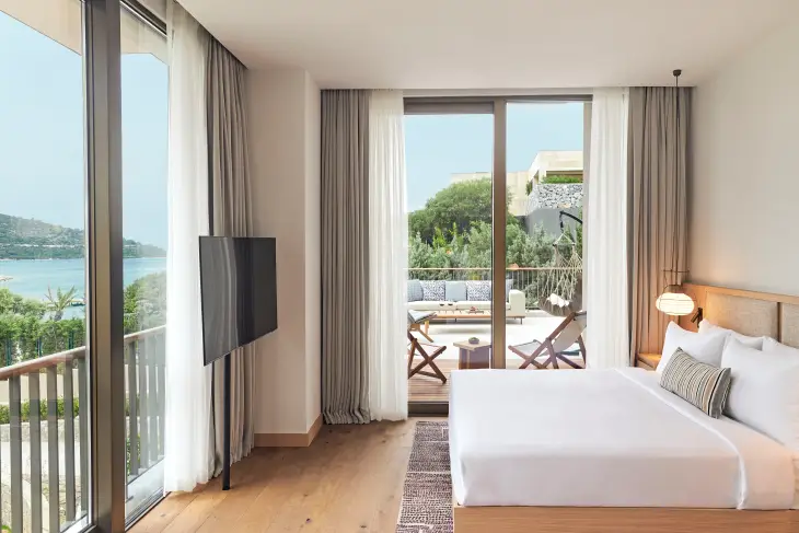 Bodrum'un en iyi otellerinden birinde, denize nazır modern ve ferah bir otel odası, rahatlatıcı beyaz çarşaflı bir yatak, geniş pencerelerden görünen masmavi deniz manzarası ve konforlu dış mekan mobilyalarıyla süslenmiş özel balkonuyla huzurlu bir tatil vaat ediyor.