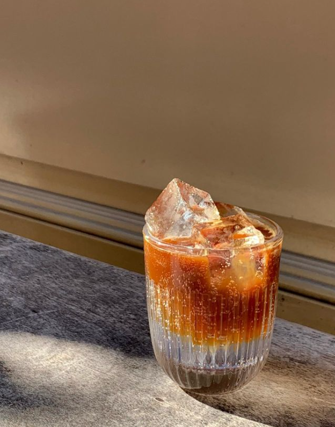 Güneşli bir Paris restoranında ahşap bir masada duran, dibi şeffaf gazlı içecek ve üstü koyu amber rengi kahve veya çaydan oluşan, içinde büyük buz küpleri olan şık, nervürlü bir bardak, şehrin keyifli gastronomi deneyimlerinden birini yansıtıyor.