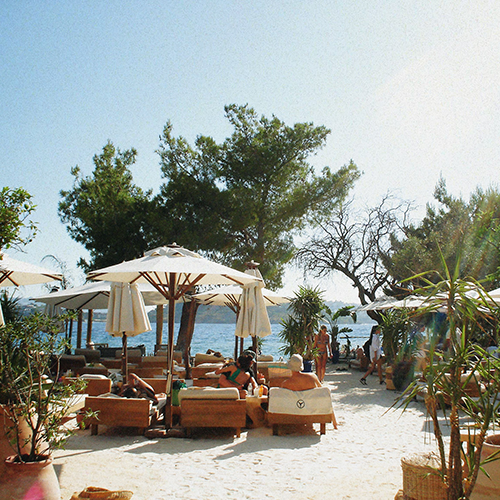Bodrum'un gözde lüks beach club'ı MOMO Bodrum'da, Ege'nin mavisinde parıldayan denize nazır uzanan bembeyaz kumlu plajda, şık beyaz şemsiyeler ve konforlu ahşap şezlonglar altında güneşlenen misafirler, doğal yeşilliklerle çevrili dingin bir yaz gününün tadını çıkarıyor.