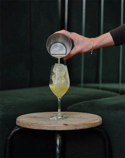 Koyu yeşil, şık bir fonda, buzlu kadehe metal shakerdan köpüklü ve açık renkli bir içecek dolduran barmen, En İyi Paris Restoranları listesindeki modern kokteyl ve şarap barlarının dinamik atmosferini yansıtıyor.