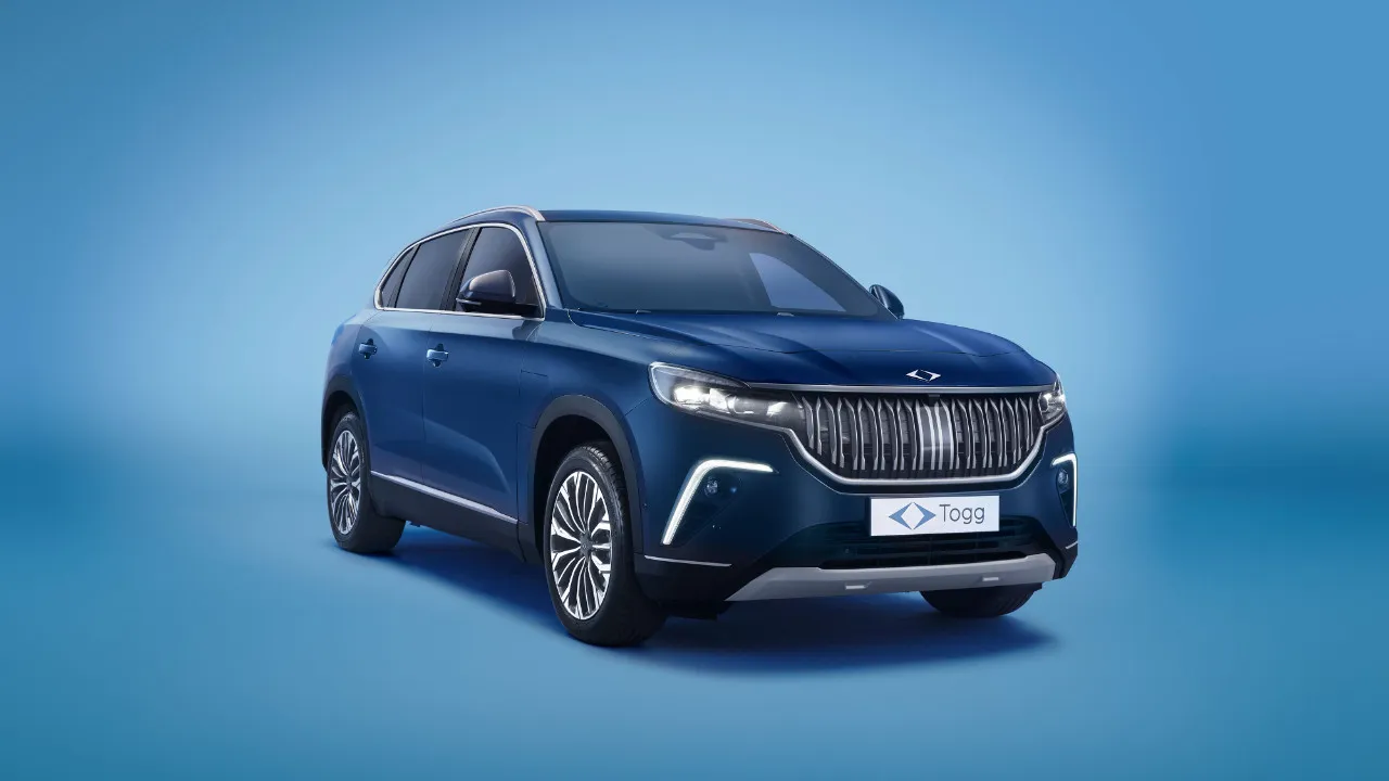 Türkiye’de Yılın Otomobili ödülünün 2024 yılındaki sahibi olan, parlak lacivert Togg T10X SUV modeli, dinamik ön farları ve modern ızgara tasarımıyla dikkat çekerek, açık mavi tonlardaki bir stüdyo ortamında ön cepheden çapraz duruşla sergileniyor.