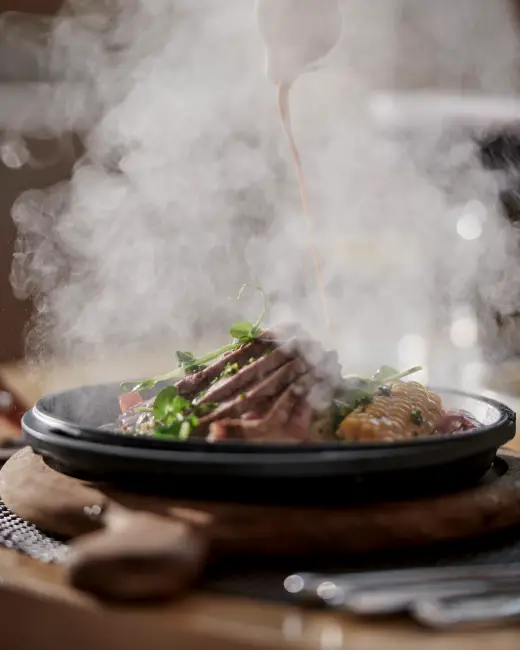 Bodrum'un en iyi restoranları arasında yer alan Voyage Torba'nın Gastro Street bölümünde sunulan, közlenmiş mısır ve taze yeşilliklerle zenginleştirilmiş dilimlenmiş etin üzerine buharla birlikte lezzetli bir sosun döküldüğü anı gösteren nefis bir yemek tabağı.