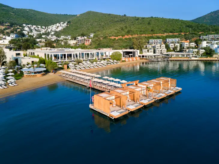 Bodrum'un Torba bölgesindeki lüks bir otelin kuşbakışı görüntüsünde, uzun kumlu plajında sıralanmış beyaz şemsiyeler ve şezlonglar, denize uzanan iskele üzerindeki ahşap özel kabinler ve yeşil tepelerle çevrili tesisin sunduğu ayrıcalıklı tatil imkanı göz alıcı.
