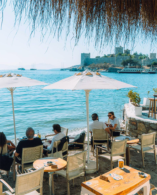 Bodrum'un en iyi restoranları arasında yer alan Yula'da, masmavi Ege Denizi kıyısında, tarihi Bodrum Kalesi manzarasına karşı güneşli bir günde içeceklerini yudumlayan insanlar görülüyor.