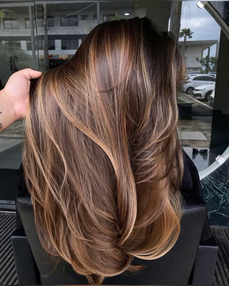 Rich Girl Brunette saç trendinin yansıdığı bu görselde, sırtı dönük bir kadının omuzlarına dökülen, parlak, sağlıklı ve eforsuz dalgalı saçları, zengin kahve tonları ve ışıltılı sarı balyajlarla lüks bir görünüm sunuyor.