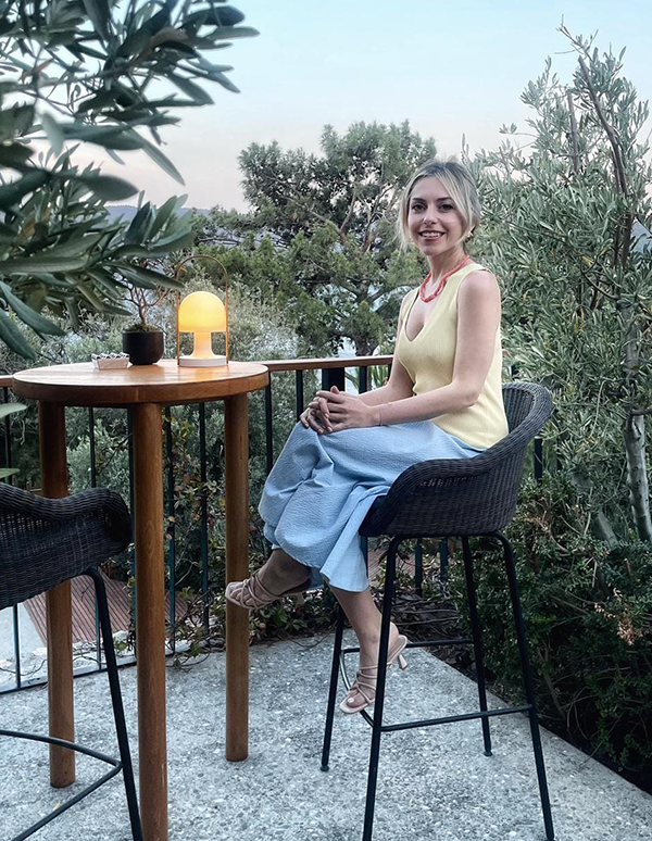Bodrum'un en iyi restoranlarından biri olan Loft Elia'nın zeytin ağaçları arasındaki açık hava masasında, sarı bluzu ve açık mavi pantolonuyla oturan Pelin Hasçalık, gülümseyerek kameraya bakıyor, masasında yanan şık bir lamba ve yeşil bitkilerle çevrili akşamüstü ambiyansı dikkat çekiyor.