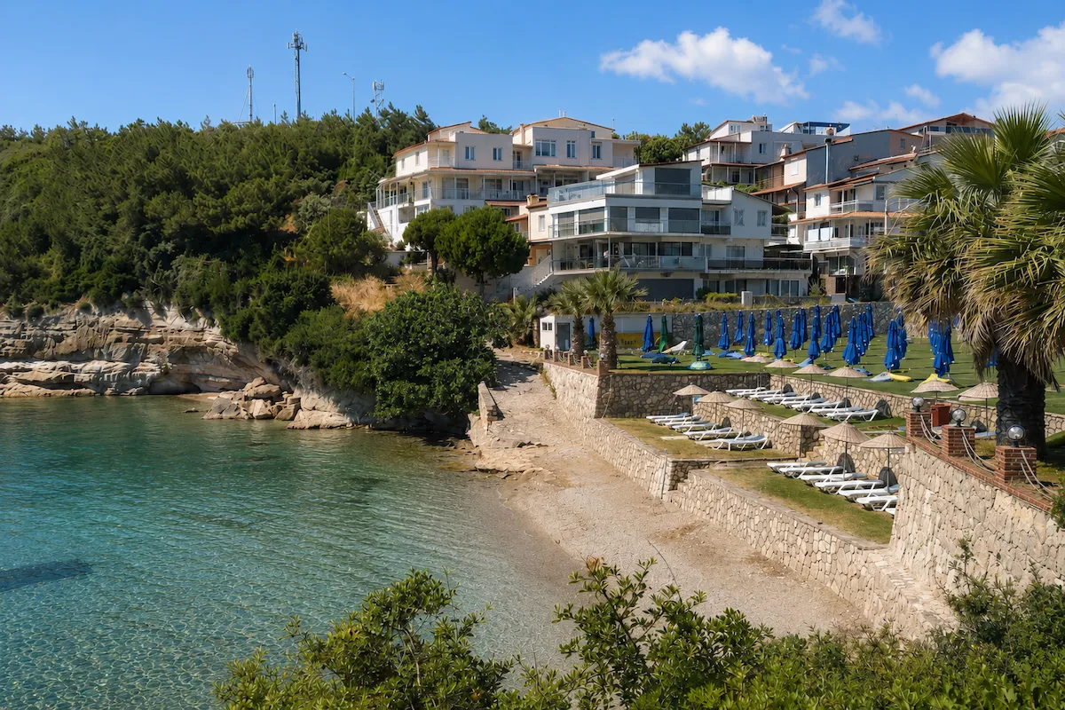 Türkiz mavisi berrak suları, dar çakıllı plajı ve yemyeşil ağaçlarla çevrili yamaca inşa edilmiş çok katlı beyaz binaların oluşturduğu Ayı Balığı Koyu, Karaburun, İzmir'de şezlonglar ve hasır şemsiyelerle düzenlenmiş teraslı bir sahil alanını gözler önüne seriyor.
