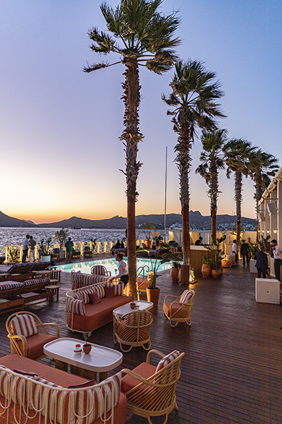 Bodrum'un Yalıkavak Marina'sında, gün batımının turuncu ve mor tonlarına bürünmüş gökyüzü altında, Fransız festive restoranı Bagatelle'in ışıklı havuz ve deniz manzaralı terasında, modern rattan oturma grupları, sıralı uzun palmiye ağaçları ve uzak tepelerle çevrili Ege Denizi, şık bir eğlence ve dinlenme atmosferi sunuyor.