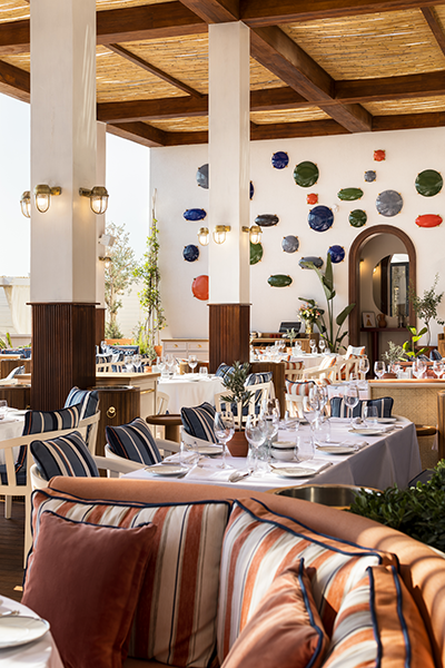 Bodrum Yalıkavak Marina'daki Bagatelle restoranının, hasır tavanı, beyaz direkleri ve renkli tabaklarla bezeli duvarlarıyla Fransız Rivierası esintisini taşıyan şık terasında, Akdeniz temalı çizgili yastıklarla süslenmiş oturma alanları ve özenle düzenlenmiş yemek masaları, Ege'nin lüks ve keyifli atmosferini vurguluyor.