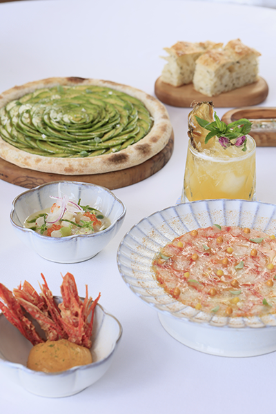 Bir masa üzerinde, avokado dilimleriyle spiral şeklinde süslenmiş bir pizza, deniz ürünlü carpaccio, kızarmış karidesler, ceviche ve ananas dilimli bir kokteyl gibi çeşitli zarif yemekler ve içecekler sergileniyor; bu sunumlar, Bodrum'un Ege'ye özgü sofistike lezzet ve eğlence anlayışını yansıtıyor.