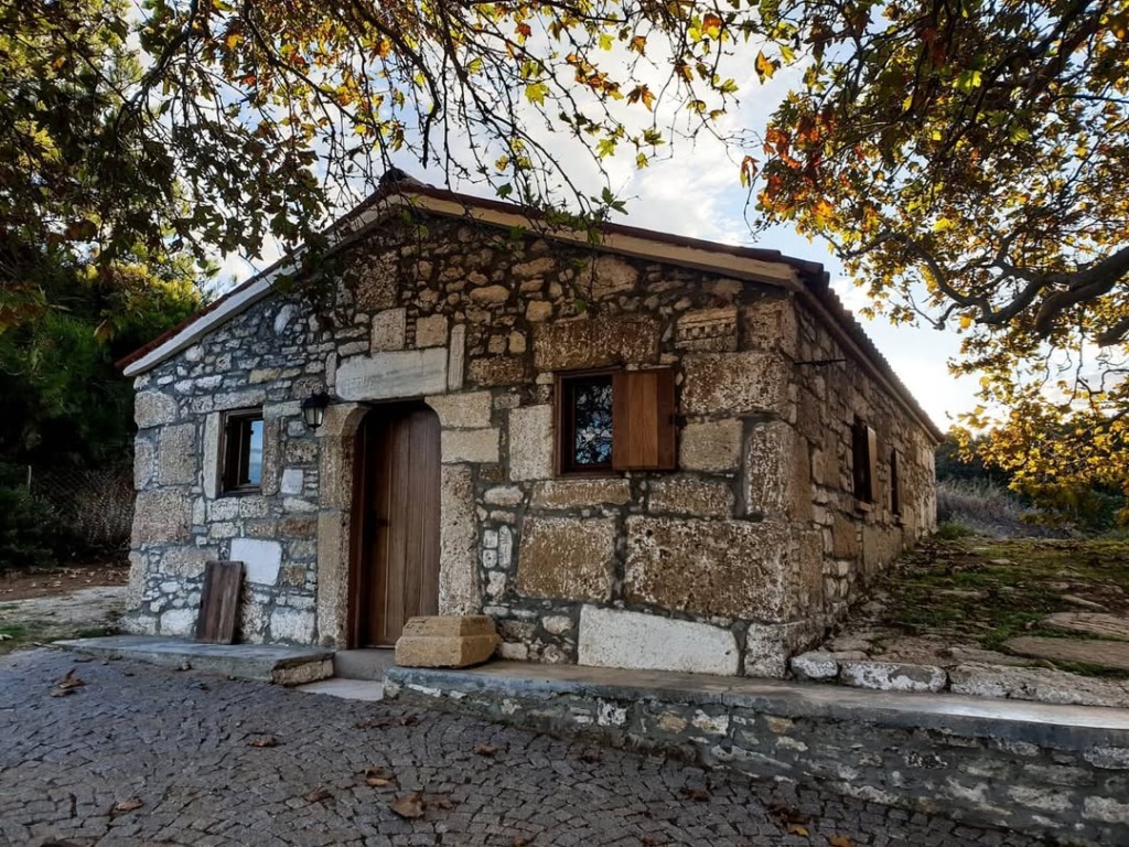 Bozcaada gezi rehberi içeriğindeki Ayazma Manastırı’nın, doğal taş duvarlara ve ahşap kapıya sahip tarihi tek katlı yapısı, sarı-yeşil yapraklı ağaçlarla çerçevelenmiş sonbahar gününde, güneş ışıklarıyla aydınlanmış ve eski bir Arnavut kaldırımlı yola bakarken görülüyor.