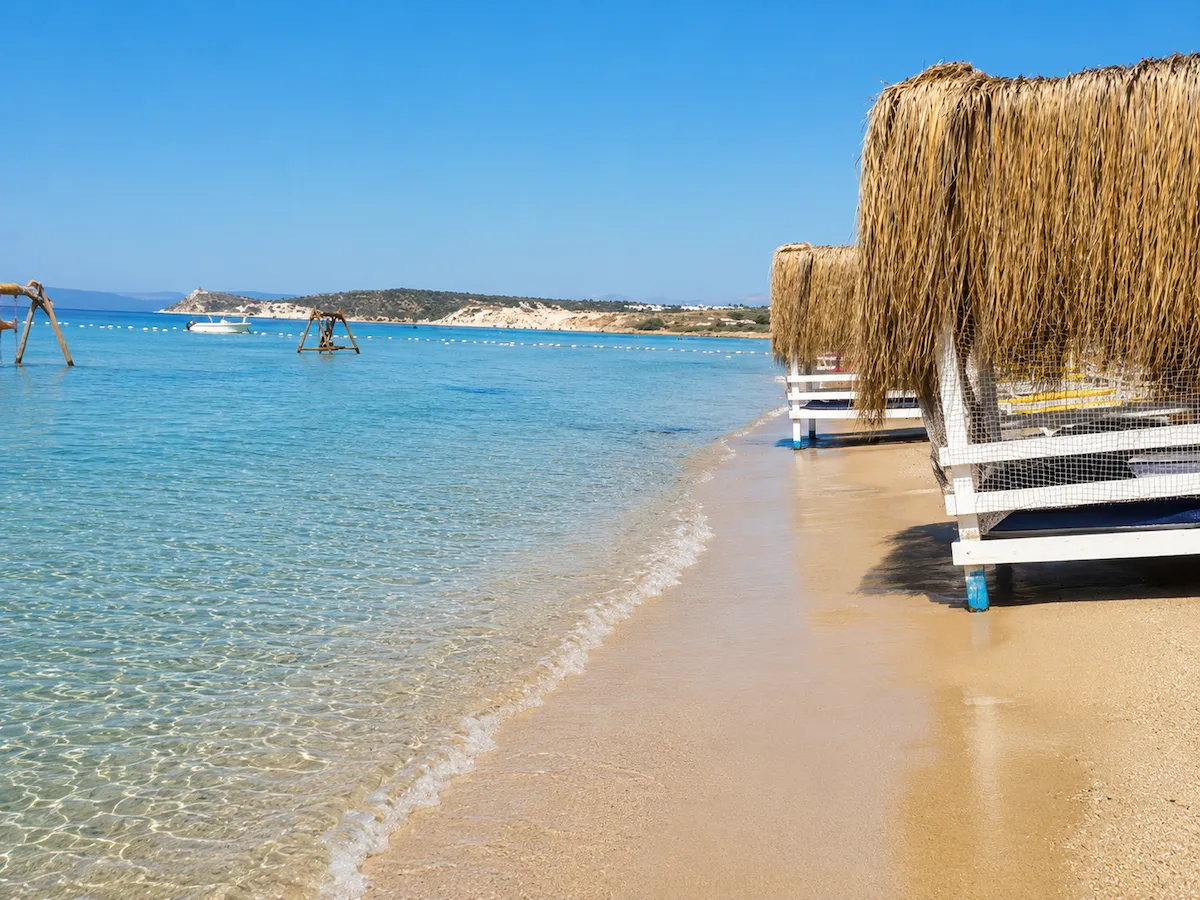 Çeşme Altınkum Plajı'nın sıcak kumlarında, masmavi gökyüzü altında berrak turkuaz denizin hafifçe dalgalandığı kıyıda, beyaz çerçeveli ve hasır çatılı modern plaj kabinleri sıralanırken, uzakta yeşil tepeler ve yüzen bir tekne görülüyor.