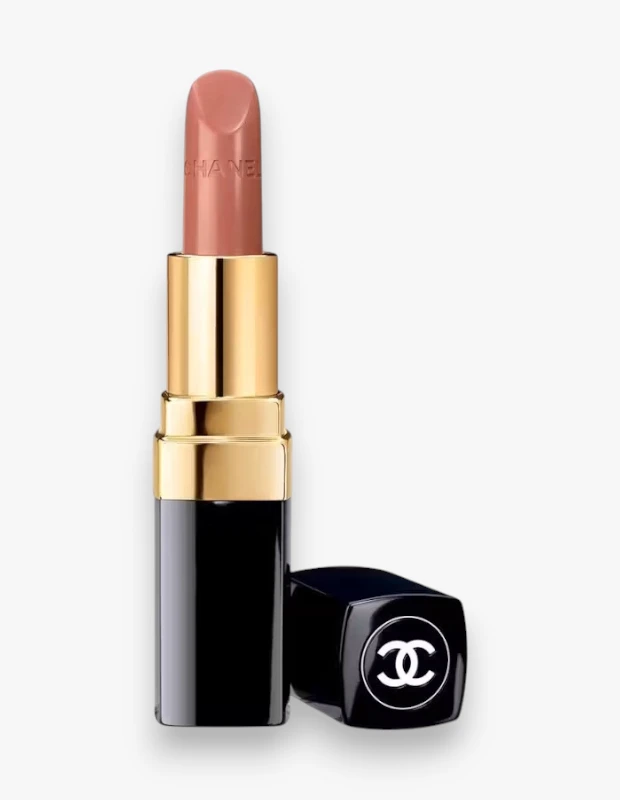 Beyaz bir arka plan üzerinde, hafifçe açılmış ve CHANEL yazısı görünen, pembe-bej tonlarında Rouge Coco Adrienne ruj, altın rengi gövde detayları ve yanında duran ikonik beyaz çift C logolu parlak siyah kapağıyla şık bir şekilde sergileniyor.