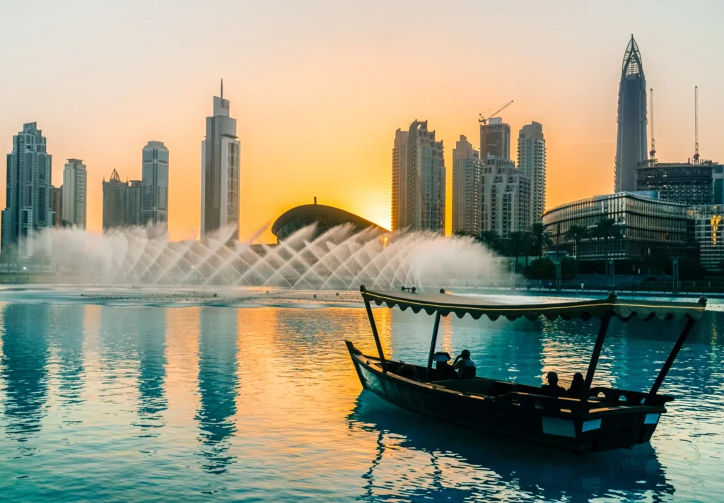 Dubai şehir silueti önünde, gün batımında su gösterileri eşliğinde ilerleyen geleneksel Arap teknesi; modern ve lüks atmosferde egzotik bir destinasyon deneyimi.