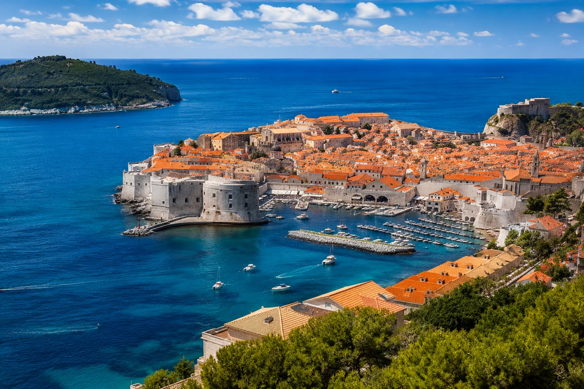 Hırvatistan'ın Dubrovnik Eski Şehri'nin panoramik bir görüntüsü, tarihi surlarla çevrili, kızıl kiremitli çatılara sahip binaların masmavi denizin kıyısında, teknelerle dolu bir limanı ve yemyeşil bir adayı içeren etkileyici bir manzarayı gözler önüne seriyor.