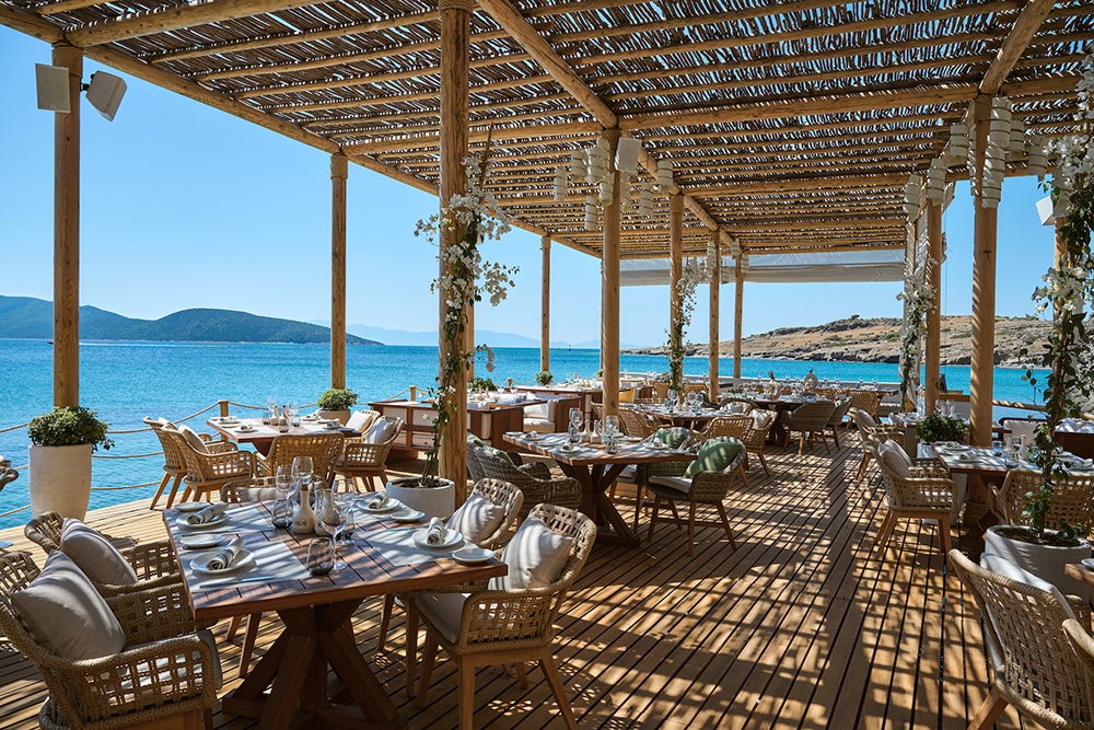 Bodrum'da, masmavi Ege Denizi'ne nazır, bembeyaz begonvillerle süslenmiş ahşap bir terasta yer alan şık bir restoran veya beach club, gölgelikli tavanı ve zarif hasır sandalyeleriyle misafirlerini Ege'nin ritmine uygun rafine bir dinlenceye davet ediyor.