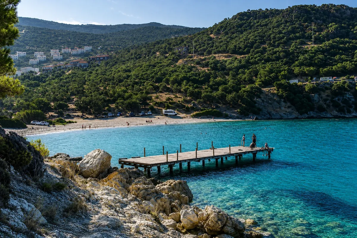 Dikili'deki Hayıtlı Koyu'nun masmavi, berrak sularında uzanan ahşap iskelenin üzerinde duran insanlar, koyun kıyısındaki kumlu plajda güneşlenen ve yüzünen ziyaretçilerle birlikte yemyeşil tepelerin eteklerindeki beyaz evlerin oluşturduğu huzurlu manzarayı tamamlıyor.