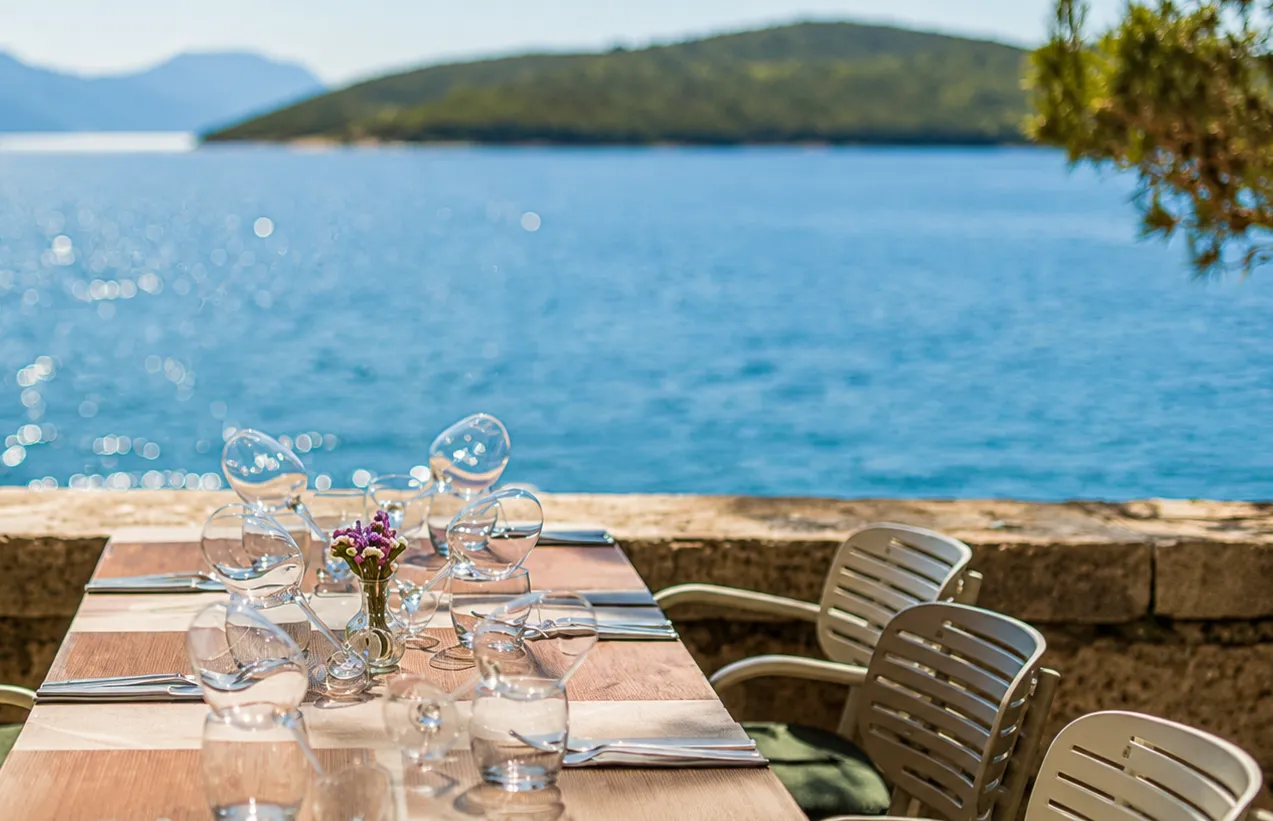 Hırvatistan'ın Korčula adasındaki Filippi restoranının denize bakan terasında, masaya özenle yerleştirilmiş şık kadeh takımları, çatal-bıçaklar ve mor çiçekler, güneşli bir günde ışıl ışıl parlayan masmavi denizin ve yeşil tepeli adanın huzurlu manzarasıyla birleşiyor.