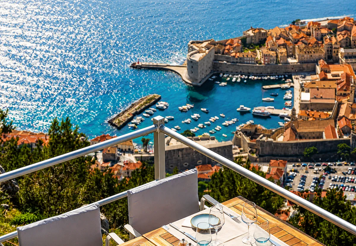 Ön planda bir masanın ve sandalyelerin bulunduğu bir panorama restorandan bakıldığında, güneşli bir günde Hırvatistan'ın Dubrovnik şehrinin tarihi surlarla çevrili limanı ve berrak mavi denizi küçük teknelerle dolu, muhteşem bir manzara sunuyor.