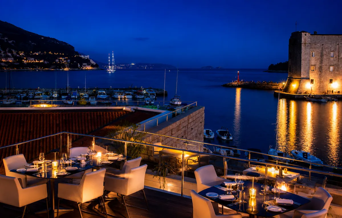 Dubrovnik'teki tarihi surların ve ışıklı limanın eşsiz gece manzarasına sahip şık bir restoran terasında, mum ışıklarıyla aydınlatılmış beyaz masa ve sandalyeler misafirlerini bekliyor.