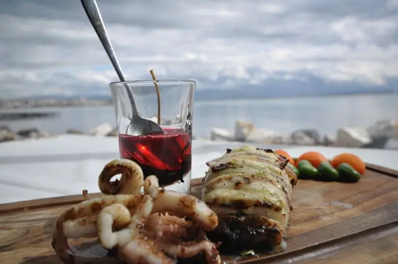Ahşap sunum tahtasında ızgara kalamar ve ahtapotun yanı sıra çeri domates ve yeşil biberlerle servis edilmiş, bir bardak kırmızı içecek eşliğinde deniz manzaralı İstasyon Restaurant'ta çekilmiş iştah açıcı bir yemek görüntüsü.