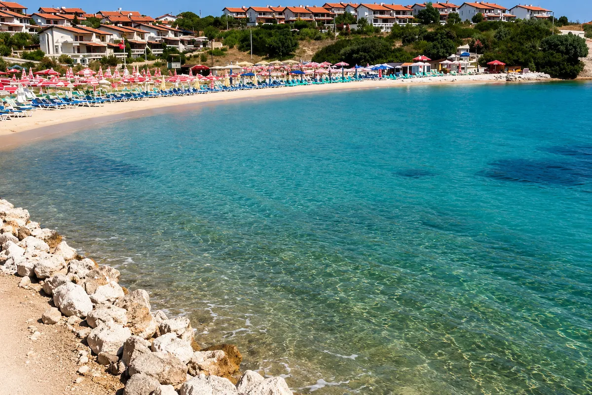 İzmir'in Dalyan bölgesindeki Ayayorgi Koyu'nun pırıl pırıl turkuaz sularıyla çevrili kumsalında, sıraya dizilmiş rengarenk şemsiyeler ve şezlonglar yer alıyor, arka planda ise yamaca sıralanmış bembeyaz evler ve yeşil bitki örtüsü manzarayı tamamlıyor.