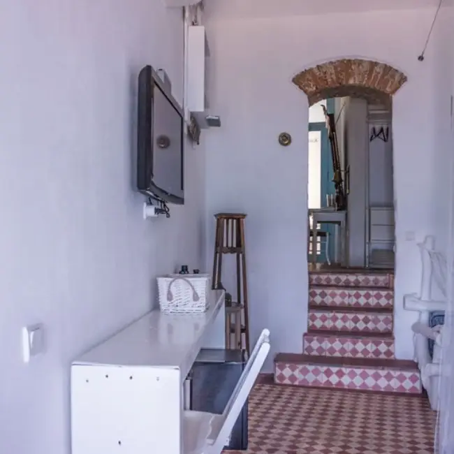 Kaikias Bozcaada'da, beyaz duvarlı, duvara monte televizyonlu bir odanın geleneksel taş kemerli girişinden görünen, pembe ve beyaz karo basamaklı bir merdiven, adanın mimari dokusunu ve Bozcaada'nın romantik kaçış rotası atmosferini yansıtan sade ve zarif bir iç mekanı sergiliyor.