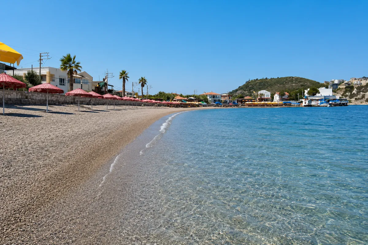 Bu parlak güneşli günde, berrak turkuaz suları çakıllı kıyıya vuran Karaburun Bodrum Koyu Plajı'nda, sıralanmış kırmızı şemsiyeler ve palmiye ağaçları arasında uzanan binalar, yemyeşil bir tepenin eteklerine kadar uzanıyor.