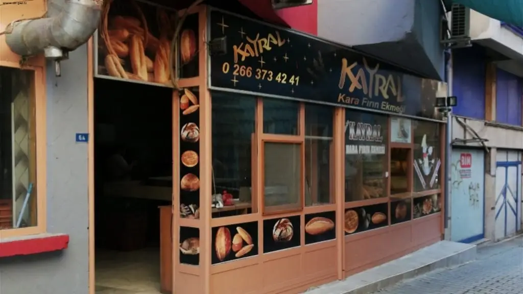 Kayral Kara Fırın Ekmeği tabelası, 0 266 373 42 41 telefon numarası ve dış cephesinde çeşitli ekmek çeşitlerinin görsellerinin sergilendiği, geleneksel ahşap çerçeveli cam vitrinlere sahip bir ekmek fırını görülmektedir.